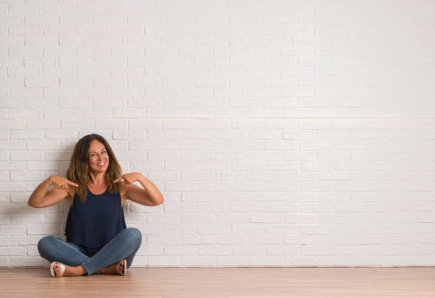 istock Middle age hispanic woman sitting on the floor over white brick wall looking confident with smile on face, pointing oneself with fingers proud and happy. 1044763808