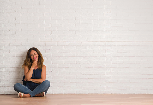 Middle age hispanic woman sitting on the floor over white brick wall looking confident at the camera with smile with crossed arms and hand raised on chin. Thinking positive.