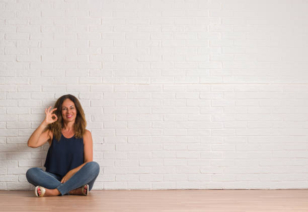istock Middle age hispanic woman sitting on the floor over white brick wall smiling positive doing ok sign with hand and fingers. Successful expression. 1044763888