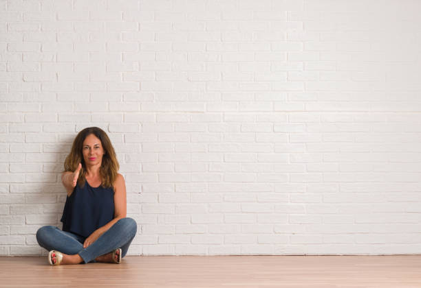 istock Middle age hispanic woman sitting on the floor over white brick wall smiling friendly offering handshake as greeting and welcoming. Successful business. 1044764096