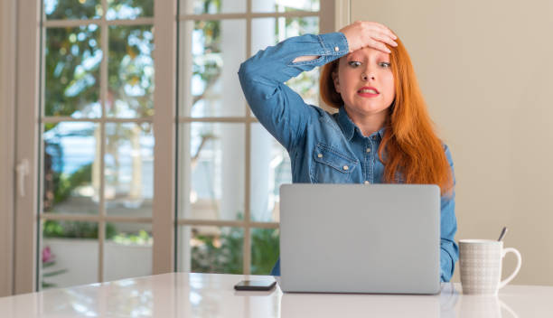 istock Redhead woman using computer laptop at home stressed with hand on head, shocked with shame and surprise face, angry and frustrated. Fear and upset for mistake. 1044765110