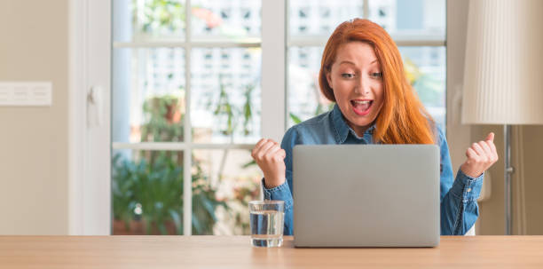 istock Redhead woman using computer laptop at home screaming proud and celebrating victory and success very excited, cheering emotion 1044767578