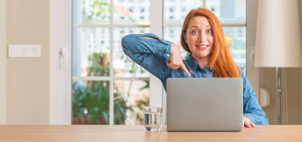 istock Redhead woman using computer laptop at home with surprise face pointing finger to himself 1044767666