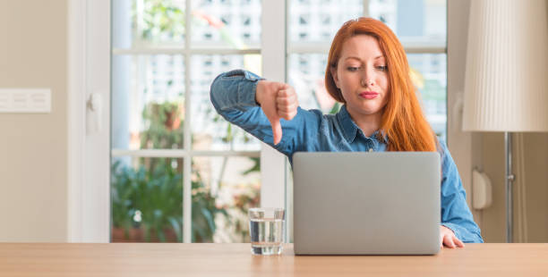 istock Redhead woman using computer laptop at home with angry face, negative sign showing dislike with thumbs down, rejection concept 1044767682