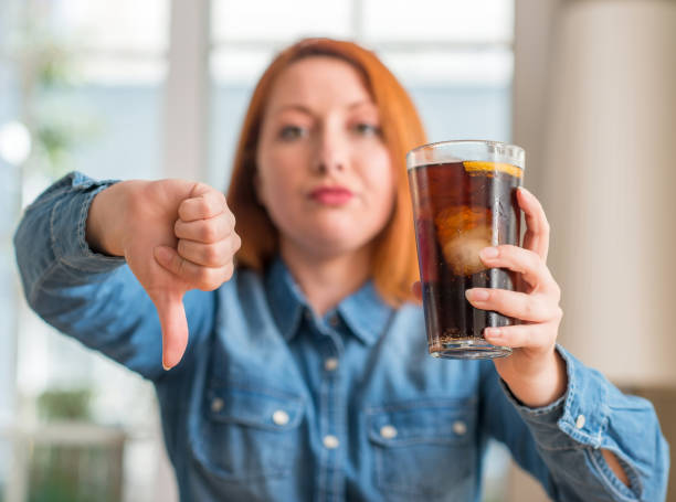 femme rousse tenant en rafraîchissement soude avec visage en colère, signe négatif montrant n’aime pas avec les pouces vers le bas, notion de rejet - dieting food rejection women photos et images de collection