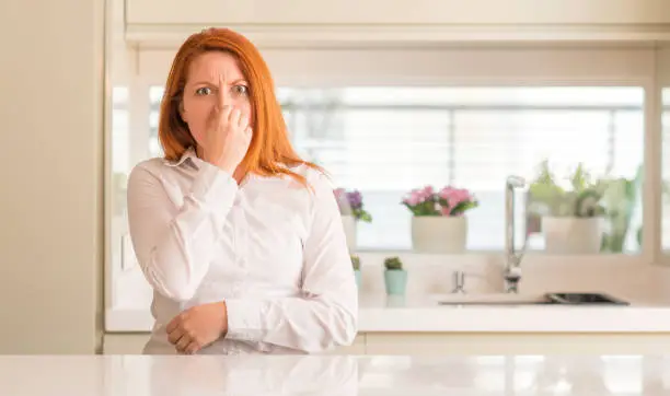 Photo of Redhead woman at kitchen smelling something stinky and disgusting, intolerable smell, holding breath with fingers on nose. Bad smells concept.
