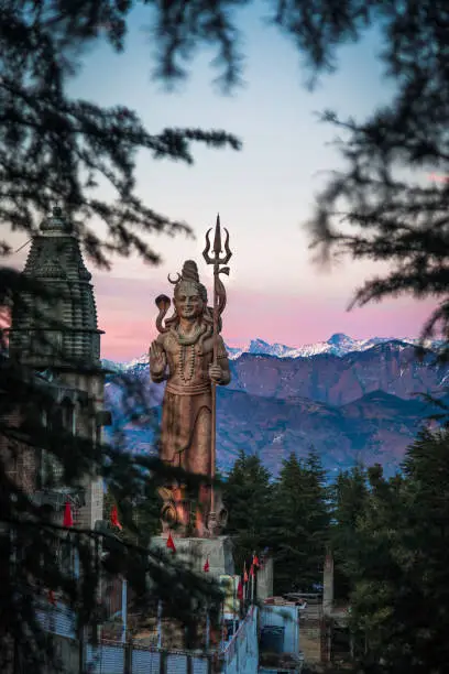 The tall golden Shiva staute of Khajjiar during the sunset with snow peaks in the background and peeking through the trees