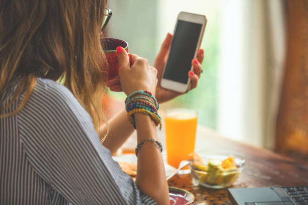 Woman using cellphone and laptop inside house and drinking coffee / tea.
