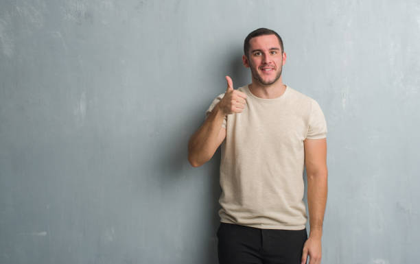 istock Young caucasian man over grey grunge wall doing happy thumbs up gesture with hand. Approving expression looking at the camera with showing success. 1044791178