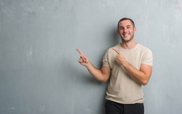 istock Young caucasian man over grey grunge wall smiling and looking at the camera pointing with two hands and fingers to the side. 1044791624