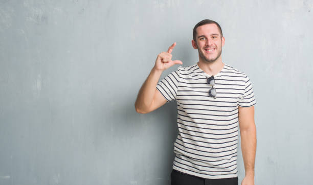 istock Young caucasian man over grey grunge wall smiling and confident gesturing with hand doing size sign with fingers while looking and the camera. Measure concept. 1044799476