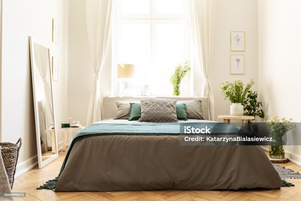 Beige, green and gray bedroom interior in a tenement house with a bed against a sunny window and bunches of wild flowers. Real photo. Bed - Furniture Stock Photo