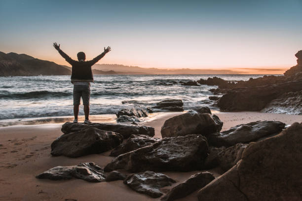 istock Young man with arms raised by the sea at sunset 1044808026
