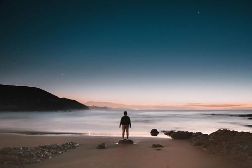 Rear view silhouette of a young man standing at an empty beach at night under a starry sky