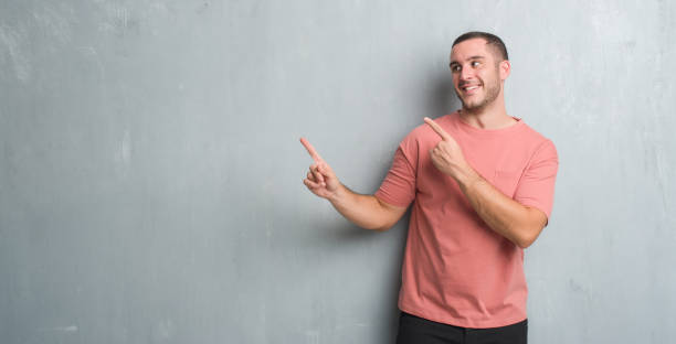istock Young caucasian man over grey grunge wall smiling and looking at the camera pointing with two hands and fingers to the side. 1044811906