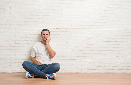 Young caucasian man sitting on the floor over white brick wall touching mouth with hand with painful expression because of toothache or dental illness on teeth. Dentist concept.