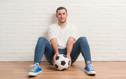 Young caucasian man sitting over white brick wall holding soccer football ball with a confident expression on smart face thinking serious