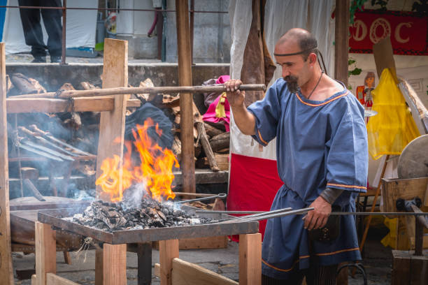 A big and strong bearded man in the protective apron is forging red hot metal rod with large forge hammer. The blacksmith is screaming looking on the hammer with a flame. Shooting at real smithy