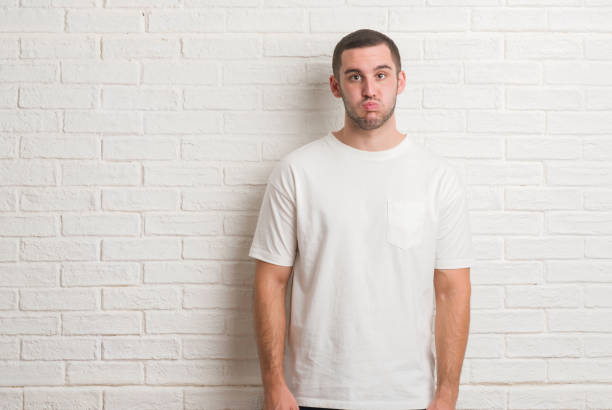 istock Young caucasian man standing over white brick wall doing ok gesture with hand smiling, eye looking through fingers with happy face. 1044824414