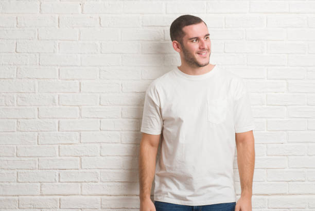 istock Young caucasian man standing over white brick wall looking away to side with smile on face, natural expression. Laughing confident. 1044824726