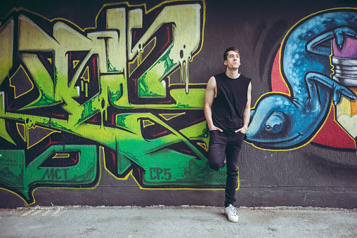 Young man is posing leaning against the wall covered with colorful graffiti art.