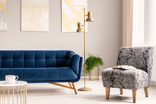 Minimal style living room interior with a navy blue couch, a patterned gray armchair and gold accents of a lamp, a coffee table and a flower stand. Real photo.