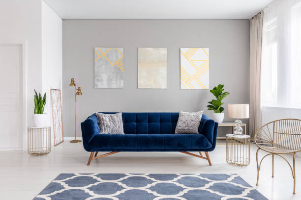 An elegant navy blue sofa in the middle of a bright living room interior with gold metal side tables and three paintings on a gray wall. Real photo. An elegant navy blue sofa in the middle of a bright living room interior with gold metal side tables and three paintings on a gray wall. Real photo. group of objects photos stock pictures, royalty-free photos & images