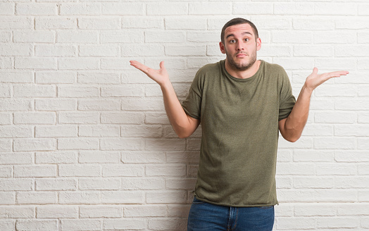 Young caucasian man standing over white brick wall clueless and confused expression with arms and hands raised. Doubt concept.