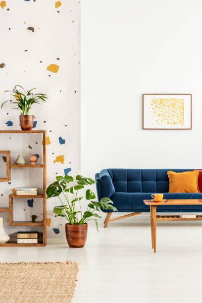istock Plants in white apartment interior with poster above blue settee with orange pillow and table. Real photo 1044829262