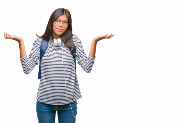 young asian student woman wearing headphones and backpack over isolated background clueless and confused expression with arms and hands raised. doubt concept. - head and shoulders audio imagens e fotografias de stock