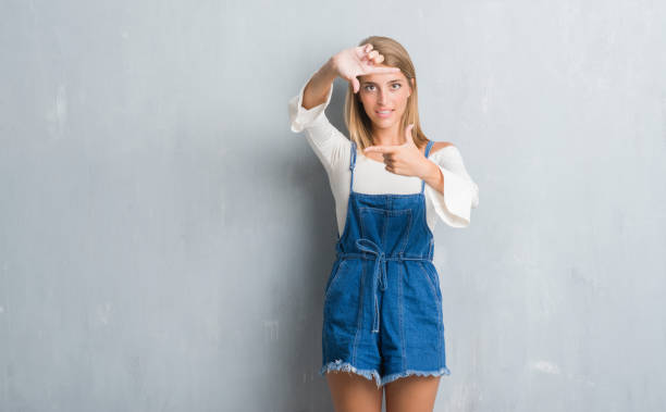 istock Beautiful young woman standing over grunge grey wall smiling making frame with hands and fingers with happy face. Creativity and photography concept. 1044859104
