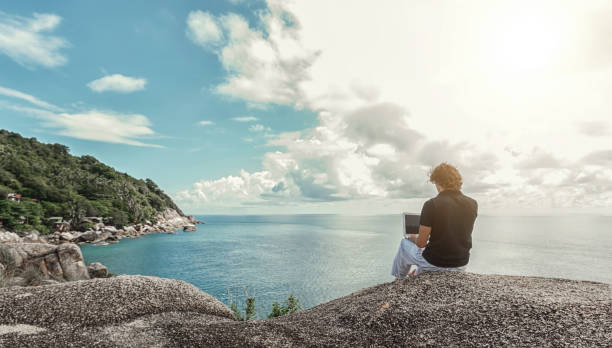 若い男が、フリーランサーの海の上の高い崖の上に座っているし、リモートで動作 - on beach laptop working ストックフォトと画像