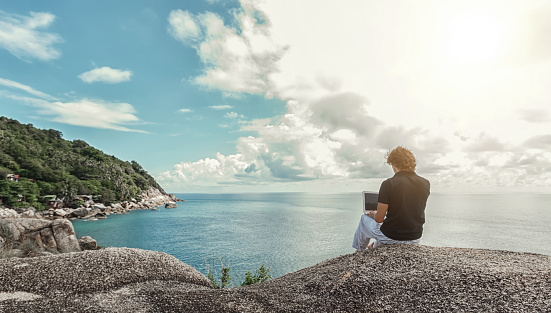 Young man, the freelancer sits on a high cliff over the sea and works remotely
