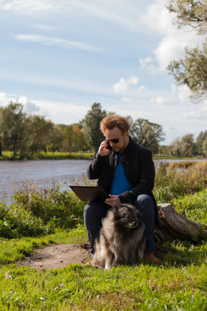 istock Young man with a laptop and a phone is sitting on a tree stump by the river 1044875162