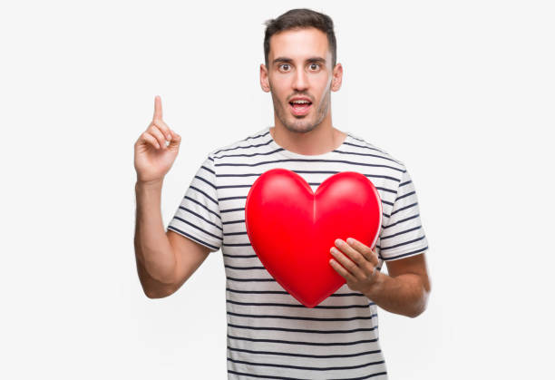 istock Handsome young man in love holding red heart surprised with an idea or question pointing finger with happy face, number one 1044883420