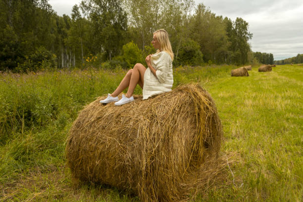istock A pretty blonde woman in a In a white knitted woolen jacket sits on a large round stack of dry hay collected on a summer or autumn day against a background of green grass at half a turn position 1044891510