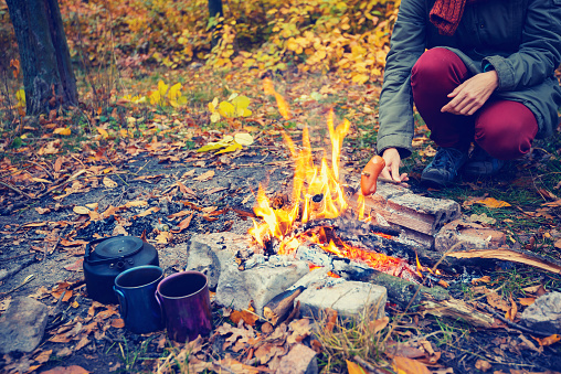 Woman roasting a sausage on a bonfire during a picnic in the autumn forest.