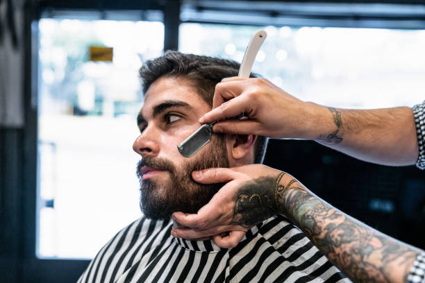 istock Close up of unrecognizable barber with tattoos on his arms shaving a customer using a razor blade 1044971506