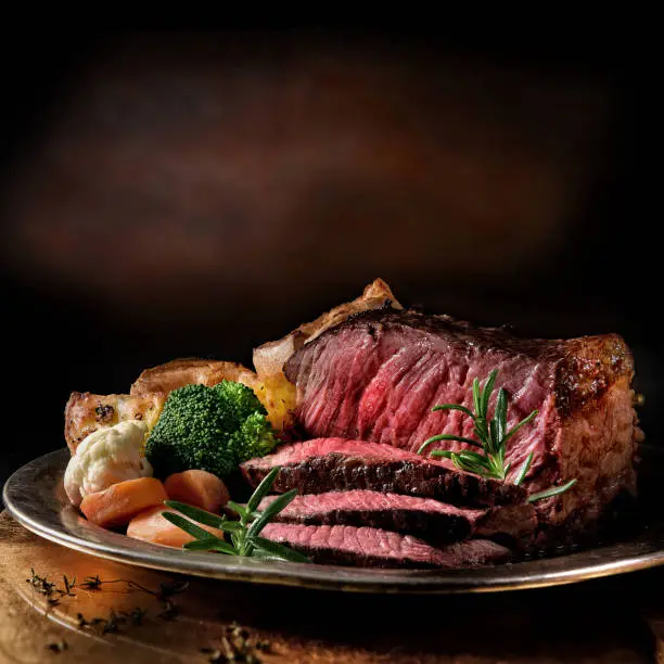 Rare roast beef meal with organic root vegetables and traditional Yorkshire pudding and roast potatoes. Shot against a dark rustic background with generous accommodation for copy space.