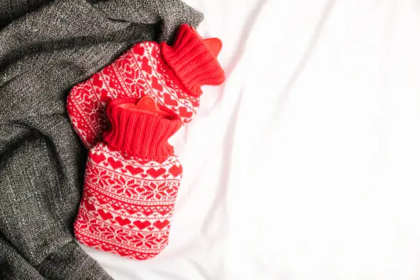 Red hot-water bottles on a bed. Heat pads on sheet, warm cozy grey knitted plaid. Top view or flat lay, copy space for your text