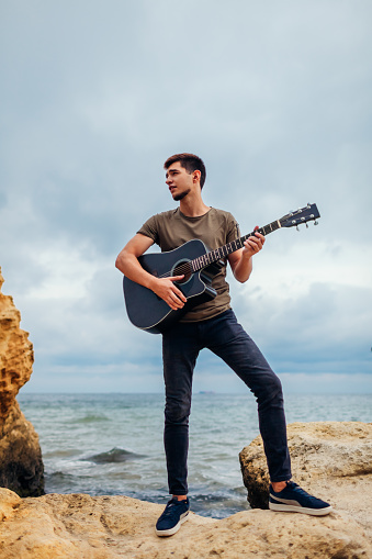 Young man with acoustic guitar playing alone on beach surrounded with rocks on rainy day