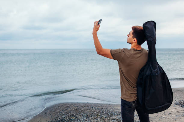 istock Young man with acoustic guitar taking selfie using phone on cloudy beach 1045039308