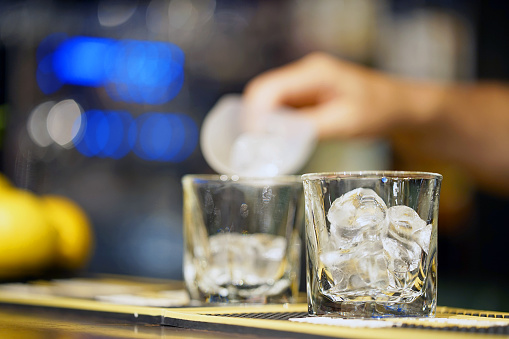Barman pours ice into a glass. Ice cubes in an empty glass, dark background, front view with details.