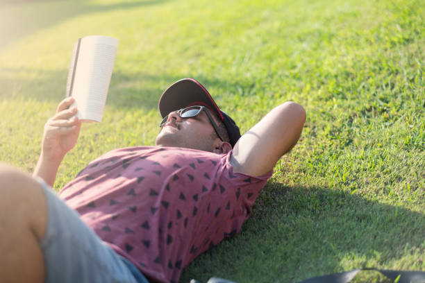 istock Young man reading a book and sitting on grass in the park 1045167944