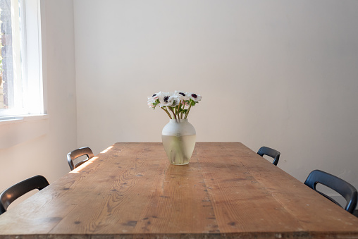 Close up of rustic wooden table with vase of white anemone flowers and four black chairs with sunlight (selective focus)