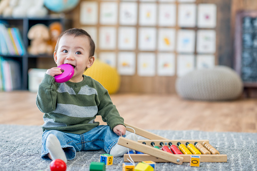 A young Asian boy is sitting on the carpet in a daycare center. He is is trying to put a toy in his mouth.