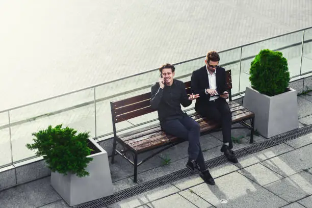 Photo of Two businessman sitting on street bench one of them is too busy by phone.