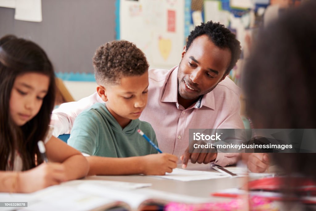 Male teacher working with elementary school boy at his desk Teacher Stock Photo