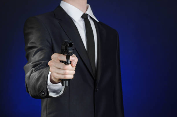 armes à feu et la sécurité thème : un homme dans un costume noir, tenant un pistolet sur un fond bleu foncé en studio isolé - gun men spy suit photos et images de collection