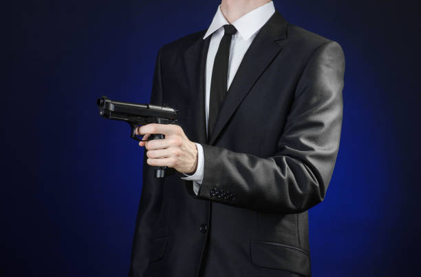 armes à feu et la sécurité thème : un homme dans un costume noir, tenant un pistolet sur un fond bleu foncé en studio isolé - handgun gun blue black photos et images de collection